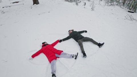 Family having fun in winter day. Young couple making snow angels. Happy man and woman lying in snow. Top view. Red coat. 4K, UHD