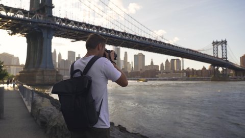 Back of young man photographer with camera, tripod setting up outside outdoors in NYC New York City Brooklyn Bridge Park by east river, railing, looking at view of cityscape skyline