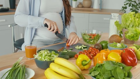 Pregnant Woman in the Kitchen Cuts Tomatoes, Prepares Dinner. Healthy, Organic and Wholesome Food. Vitamins From Fresh Vegetables for the Mother Of ohe Unborn Child. Prepare a Homemade Meal. Close-up.