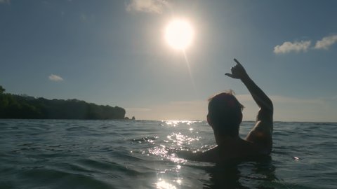The silhouette of a young man who shows the Shaka surfer gesture to the sky against the background of waves, sun and blue sky. A young surfer swims in the water and shows a surfer greeting gesture.