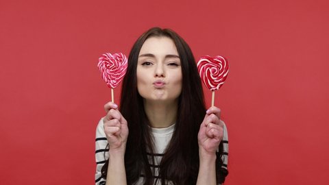 Portrait of playful woman covering eyes with heart shape lollipops, having positive expression, wearing casual style long sleeve shirt. Indoor studio shot isolated on red background.