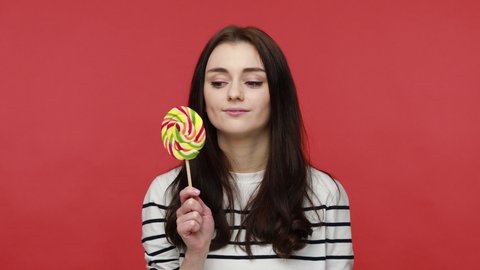 Portrait of hungry woman licking tasty lollipop, expressing happyness, enjoying delicious candy, wearing casual style long sleeve shirt. Indoor studio shot isolated on red background.