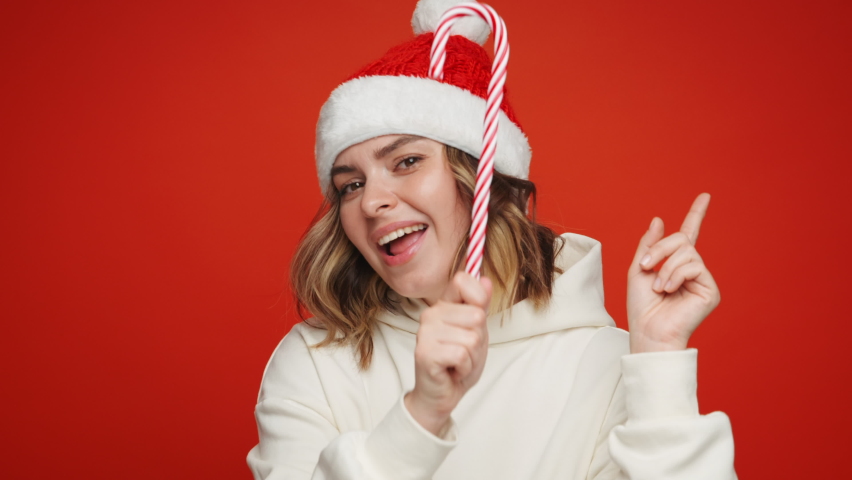 Christmas Happy New Year. Noel Happy young woman in Christmas clothes hat Santa dancing sings to music rhythmically moving her fingers with Candy smiling happily looking at camera on red background