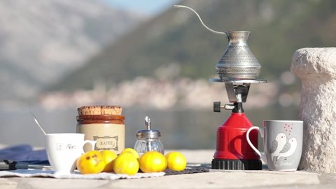 Red camp gas burner and coffee cezve outdoor on picnic at pier in bay of Donji Stoliv, Montenegro. Coffee crockery still life near ancient stone bollard on pier with mountain blurred background