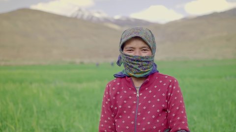 A shot of a happy young rural Asian working woman farmer with a scarf around her head and neck standing beside the green agricultural field looking at the camera and smiling 
