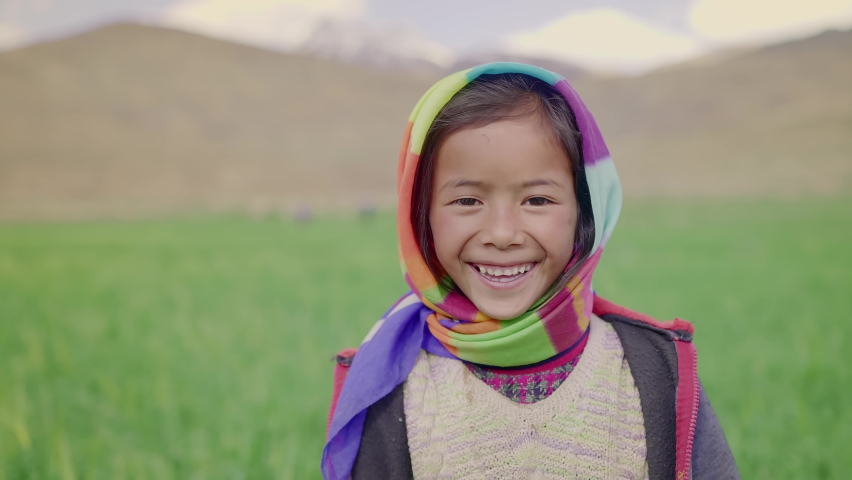 Close up shot of a happy young rural Indian Asian cute little girl with a colorful scarf around her head and neck standing beside the green agricultural field looking at the camera and smiling  Royalty-Free Stock Footage #1081177784