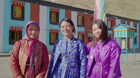 A group of three young Asian beautiful females wearing colorful traditional designer robes standing together looking at the camera smiling with Buddhist Monastery in the background.