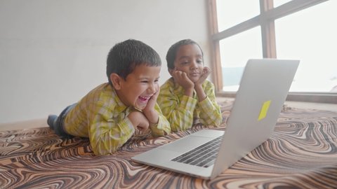 Two Indian Asian school kids wearing school uniforms are engrossed and attending online video classes using a laptop in an indoor house setup. Remote learning and education concept. 