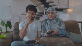 An Arabic Muslim happy couple is sitting on a couch and holding joysticks while playing a video game together. Middle eastern Husband and wife are enjoying quality time together