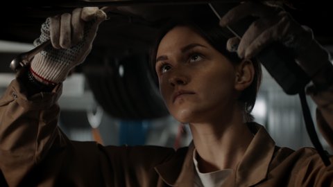 CU portrait of Caucasian female mechanic repairing a car in a workshop, working under car bottom. Shot with 2x anamorphic lens