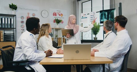 Beautiful confident qualified arabian female doctor in hijab reporting about dna structure to attantive professional skilled diverse colleagues during meeting in clinic office