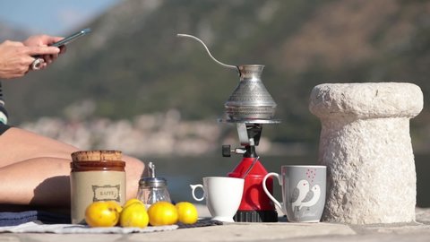 Female hands with smartphone and red camp gas burner with coffee cezve on picnic at pier in bay of Donji Stoliv, Montenegro. Coffee still life near stone bollard with blurry mountain background