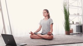 Yoga practice. Home training. Video lesson. Teenager girl sitting lotus pose meditating with laptop on floor light room interior motion view.