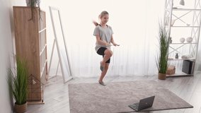 Teenager gym. Home training. Video lesson. Happy girl doing jumping workout rising high knees looking laptop light room interior.