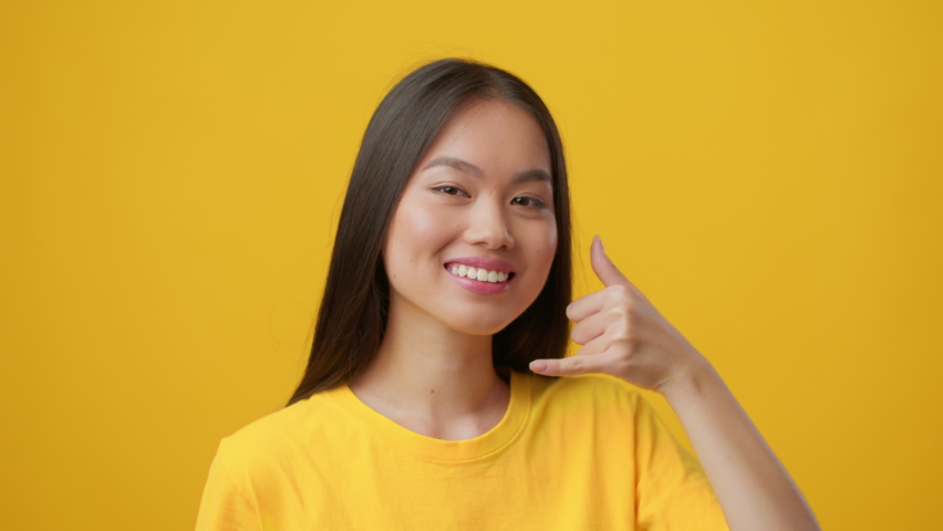 Japanese Young Woman Gesturing Call Me Holding Hand Like Cellphone Near Ear And Smiling To Camera Standing Over Yellow Studio Background. Communication Concept. Slow Motion