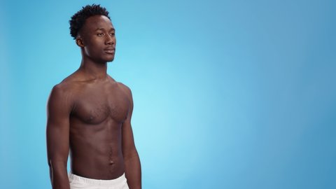 Bodybuilding concept. Young confident african american man athlete posing shirtless over blue studio background, looking aside at empty space, slow motion