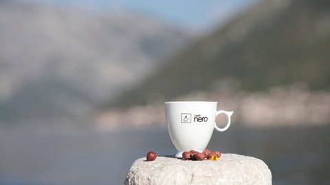 White crockery coffee cup and hazelnuts on old stone in bay of Stoliv old town. Caffe Nero brand coffee cup closeup. Rack focus from foreground to background. Donji Stoliv, Montenegro, 10 24 2021