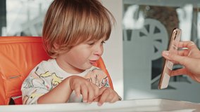 Portrait of a playful fair-haired boy, waiting for his breakfast at home. Mother shows cartoons on her smartphone to her son to grab his attention before feeding. High quality 4k footage