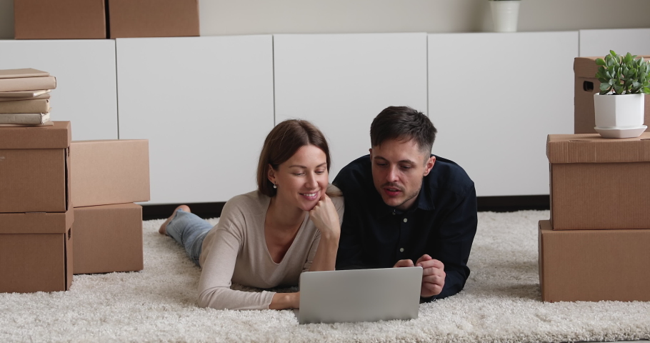 Happy loving young family couple woman and man lying on floor carpet between carton boxes, using computer applications planning interior decoration, discussing purchasing new stuff, shopping online. Royalty-Free Stock Footage #1081201361