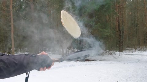 Cooking pancakes for the spring holiday Maslenitsa in the woods in early spring. Man flip pancake in the air with frying pan in slow motion