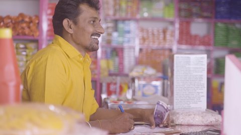 Happy Kirana Store merchant noting down orders from customers with stack of money in hand - concept of successful small businesses
