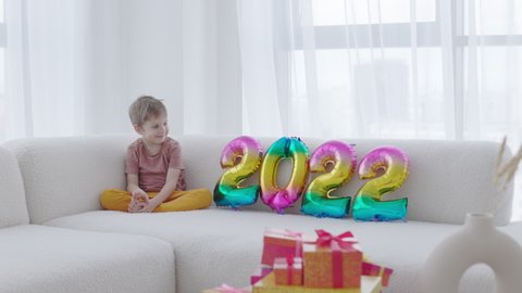The Season of Giving concept. Happy smiling kid is sitting near Christmas gift box and 2022 colorful balloons at home. Winter time, New Year presents and seasonal tradition - gift-giving.