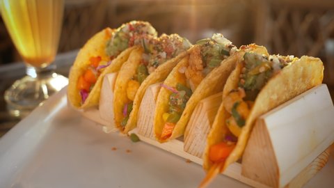 A man takes an appetizing Mexican tacos from a white plate in the rays of warm sunlight. Dinner with tacos with a bright filling and a drink, on a glass table during sunset in a cafe. Close-up.