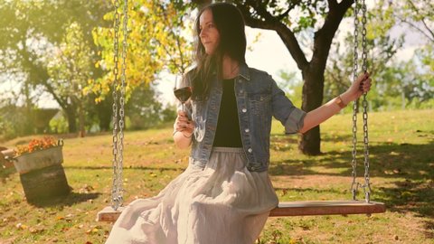 Woman sitting on swing outdoor in garden relaxing drinking wine. Sun light, sunny autumn mood, tranquility.