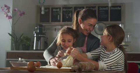 Cinematic authentic shot of happy mother and her little daughters are having fun to make dough with flour and knead it together for baking cookies or bread in kitchen at home.