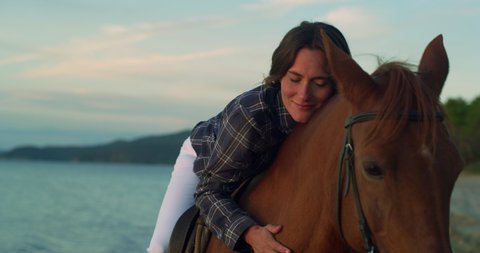 Cinematic shot of young happy woman is lying on brown purebred horse and caressing it with love and affection on seaside beach at sunset.