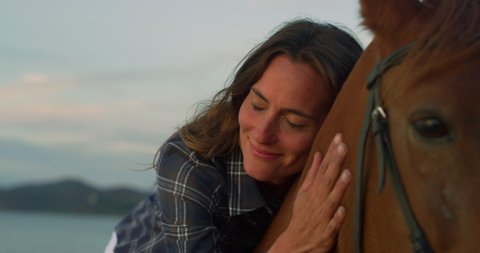Cinematic close up shot of young happy woman is lying on brown purebred horse and caressing it with love and affection on seaside beach at sunset.