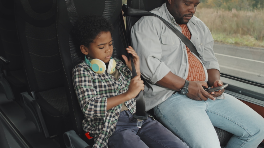 Medium shot of cute 8-year-old African-American boy in casualwear fastening seat belt while sitting together with his father in bus before ride. Man texting messages on smartphone