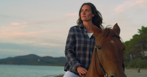 Cinematic close up shot of young happy carefree woman is riding purebred brown horse on seaside at sunset.