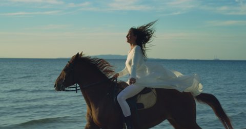 Cinematic close up shot of young happy carefree woman wearing long white dress is riding her purebred brown horse in sea at sunset.