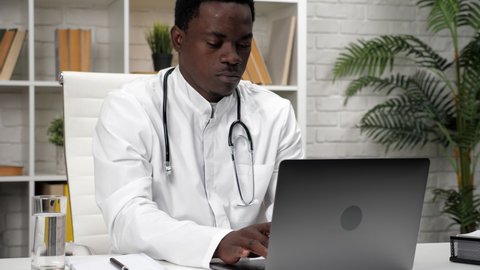 Adult African American man doctor typing text on keyboard laptop in clinic office. Medical worker in white coat working and uses computer enters patient documents into electronic database at hospital