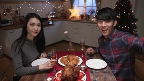 young asian couple enjoy dinner at home on xmas eve. asian man and woman face camera having video call greetings to parents online. male and female celebrate Merry Christmas of holiday near the Tree