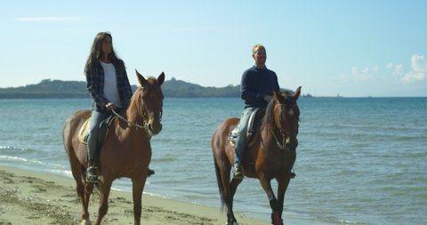 Cinematic shot of young happy carefree couple in love is enjoying to ride together purebred brown horses during romantic vacation on seashore with sun shining.