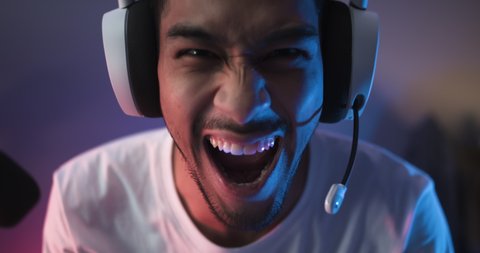 Young Asian man playing online computer video game, colorful lighting broadcast streaming live at home. Ecstatic celebration winning a match. Gamer lifestyle, E-Sport online gaming technology concept