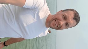 A man is talking on a video call on a smartphone against the background of the sea. A male communicates online on vacation. Vertical video.