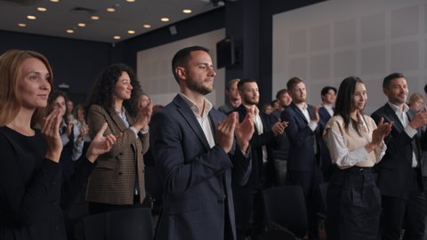 Group of people applauds to speech at business conference. Stage talk at summit meet in crowded modern hall. Caucasian man training at economy forum. Applause of corporate male person in suit indoors