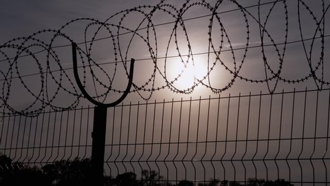 Barbed Wire Hangs on Border of an Iron Fence Against the Backdrop of Sunset. Restricted area. Barrier from strangers people, refugees, prisoners. Lattice. Jail. 4K. Close up. Slow motion.