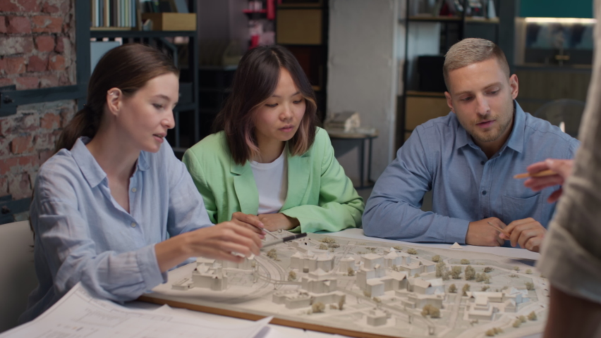 Architects and asian designer working on town area layout in modern office. Diverse group of creative people at table talking and discussing houses design. Co-workers look at model projection of homes | Shutterstock HD Video #1081232546