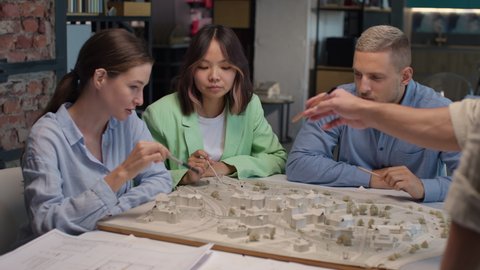 Architects and asian designer working on town area layout in modern office. Diverse group of creative people at table talking and discussing houses design. Co-workers look at model projection of homes