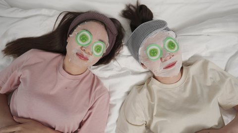 4K Smiling Asian woman friends lying on the bed with putting skin care facial mask on their face together at home. Female gay couple relax and enjoy beauty facial treatment together with happiness Video Stok