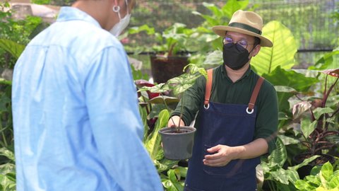 Asian man plant shop owner wearing face mask for COVID-19 pandemic protection help customer choose potted plants in greenhouse garden. Male client buying houseplant and flowers for growing at home.