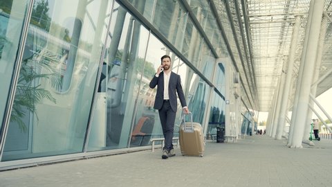 Caucasian bearded tourist businessman walks public transport building with luggage on urban background a city street. Man in formal suit trip. Business traveler pulling suitcase in airport terminal