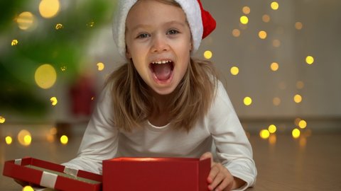 Little girl in red santa hat opens gift box with magic light surprised and laughs at dark on background of christmas tree. Happy New Year