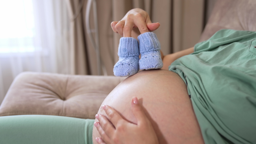 Happy awaiting caucasian lady sitting on bed and enjoying maternity, copy space. Young pregnant woman with baby booties at home | Shutterstock HD Video #1081240589