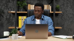 Smiling African American businessman listens online company employees laptop webcam, makes notes in notebook. Man uses computer for remote video conference call, distance business meeting in office
