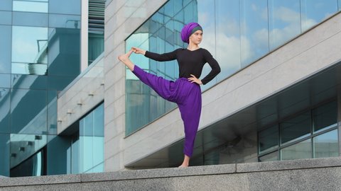 Strong muslim woman in hijab flexible young girl stretching yogi yoga workout tree pose asana female raises leg in air doing split stretch one foot balance standing city urban building background